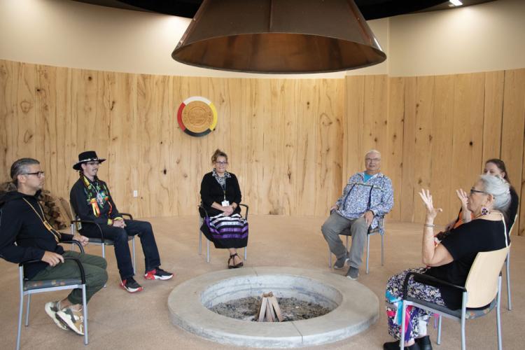 Six people sit in chairs arranged in a circle around a fire pit in a wood-paneled room