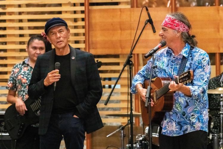 A man dressed in black next to a man singing and playing guitar 