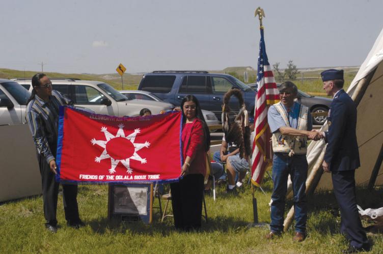 A man in civilian dress shakes hands with a man in military uniform. Two other people hold up a banner reading "Ellsworth Air Force Band: Friends of the Oglala Sioux Tribe"