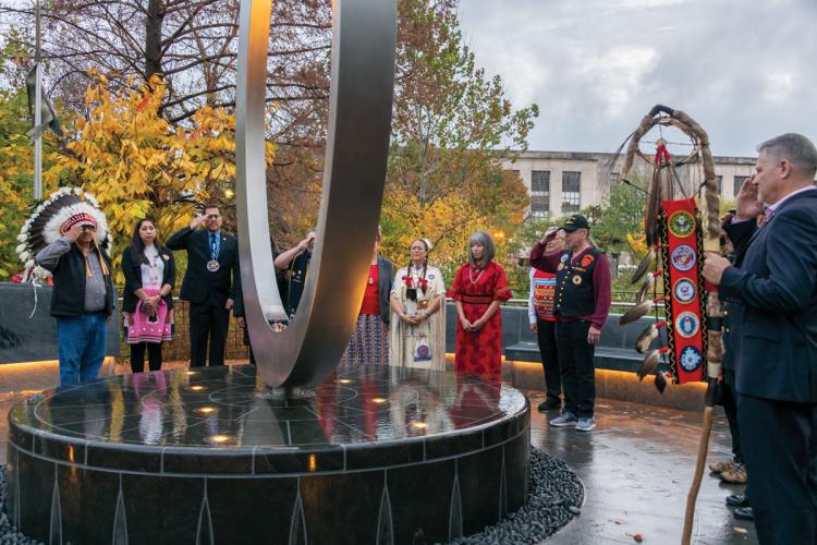 Native veterans and leaders surround a the National Native American Veterans Memorial. The veterans are saluting the ceremonial flame at its base.