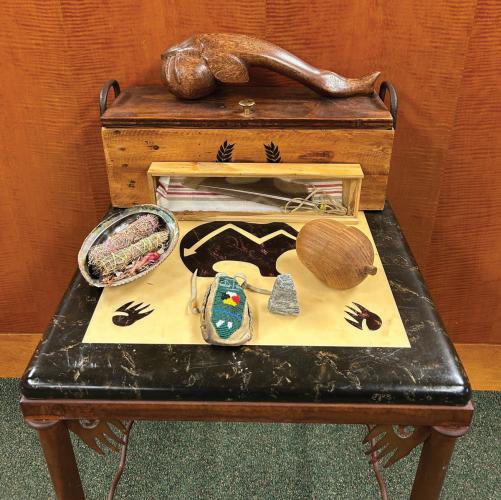 A small wood end table holding a variety of Native items, including a feather, smudge bundles, a beaded pouch, and a wood sturgeon-shaped gavel