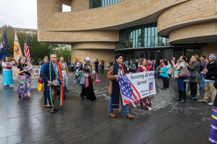 A procession of people in regalia carrying memorial banners passes by the entrance of the museum