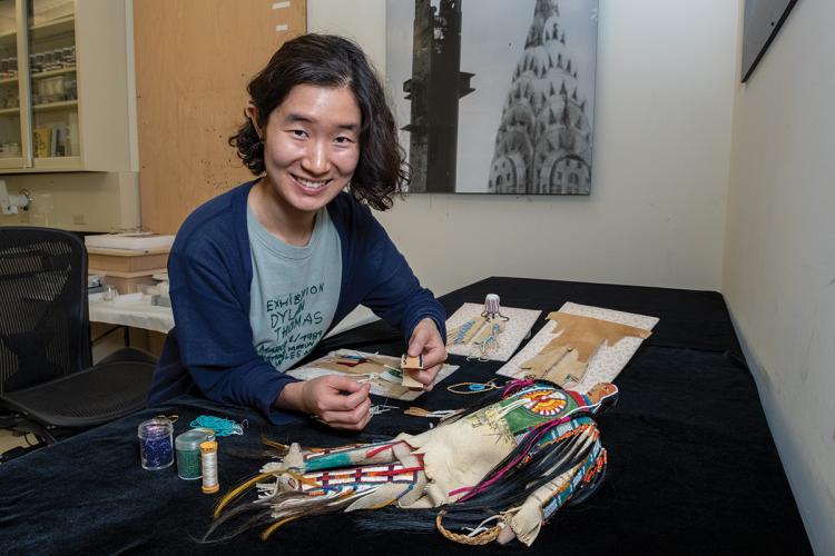 Sally Gunhee Kim holds a piece of beaded hide with a soft sculpted doll lies on a table in front of her