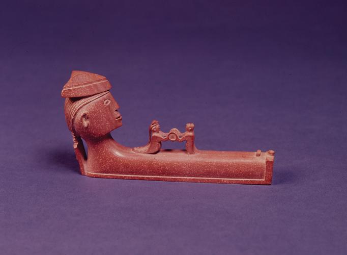 A carved brown pipe bowl depicting a face and two figures holding a whiskey barrel