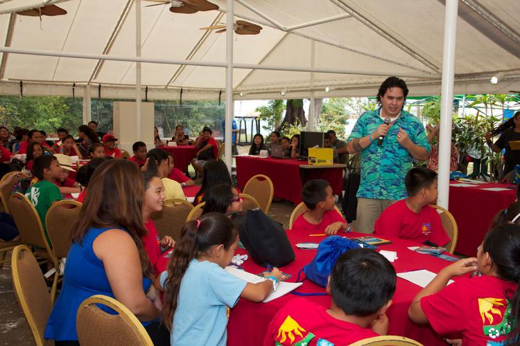 A man, standing, uses a microphone to speak to groups of seated youth
