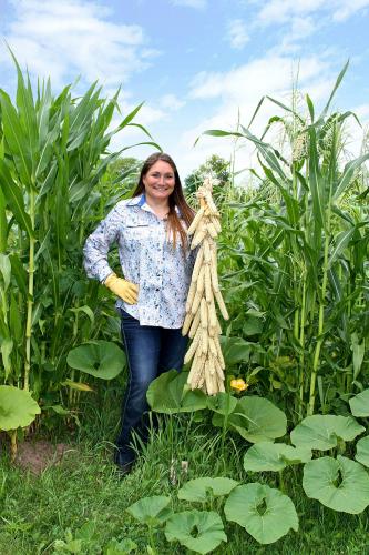 A woman stands in a field of heirloom corn, holding up a harvest of ears
