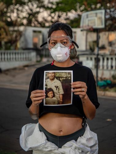 A woman wearing safety goggles and a respirator mask stands in front of the remains of her home holding a photograph