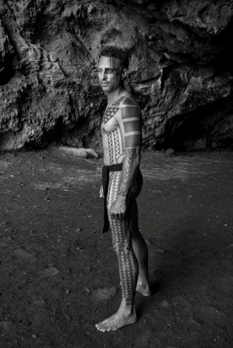 A person stands on a beach, displaying the traditional Hawaiian tattoos that cover their body