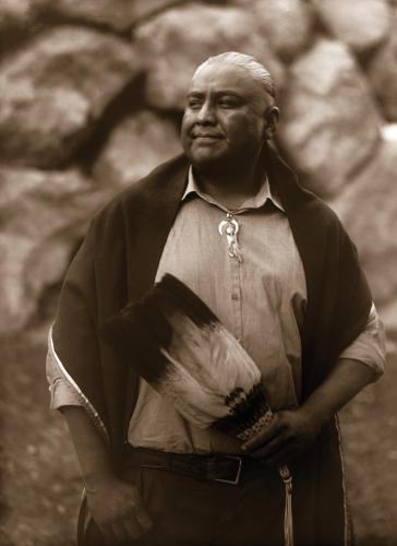 A sepia photograph of a man holding an eagle feather fan, with a blanket over his shoulders, in front of a rock wall