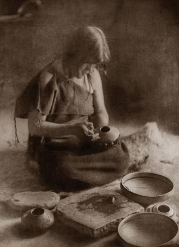 A historic photo of a potter seated cross-legged on the ground, holding a pot, with other pots around her