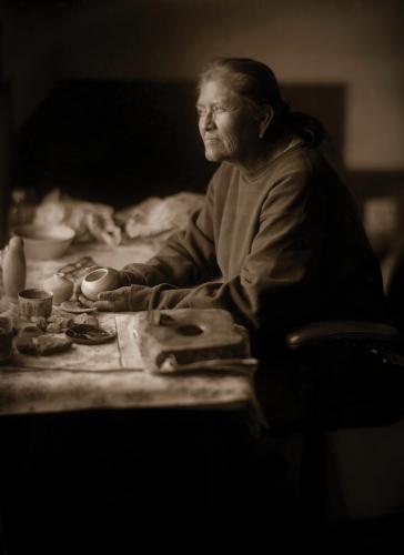 A sepia photo of a potter seated at a table, holding a small clay vessel in her hands