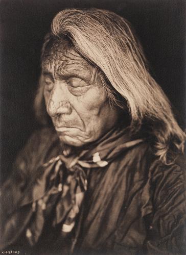 A historic photo of a Oglala Lakota elder with long grey hair, looking downwards