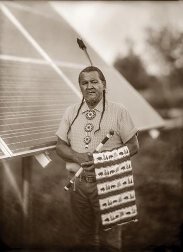 A portrait of Chief Henry Red Cloud standing in front of a solar panel holding a feather fan