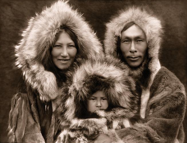 A historic photo of two Iñupiaq adults holding a young child, all wearing fur parkas with hoods