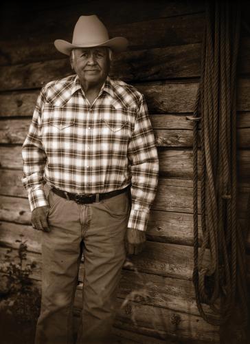 A sepia portrait of a rancher in a plaid shirt and cowboy hat, leaning against a wood wall