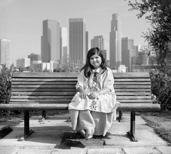 Black and white photograph of a young girl in Yalálag clothing, sitting on a park bench with the Los Angeles skyline behind her