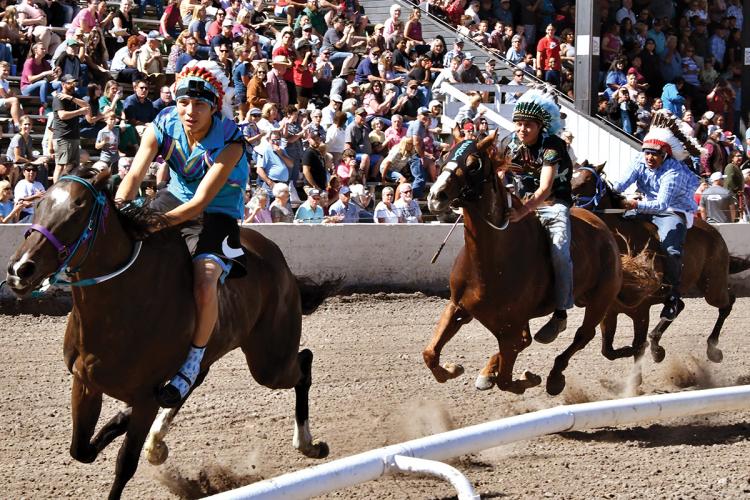 Riders wearing headdresses race horses around a track, in front of a crowd