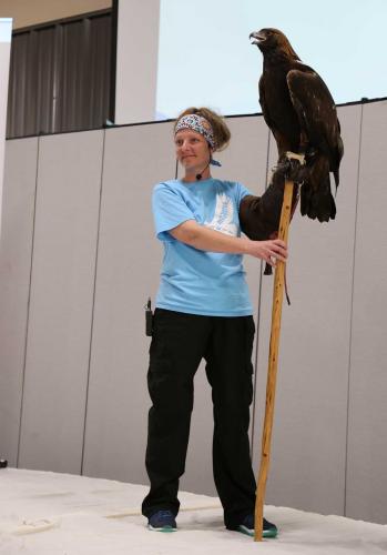A person stands beneath a presentation screen, holding a tall wooden stick on which a large golden eagle is perched