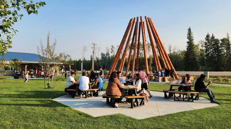 People at an outdoor pavilion near a wooden structure reminiscent of a tipi