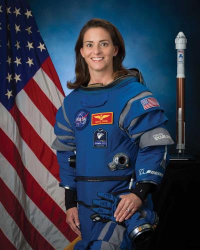 A portrait of Nicole Mann wearing a NASA space uniform, standing in front of an American flag