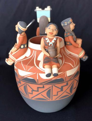 Black, red, and white patterned clay pot with four sculptures of women sitting around pot opening