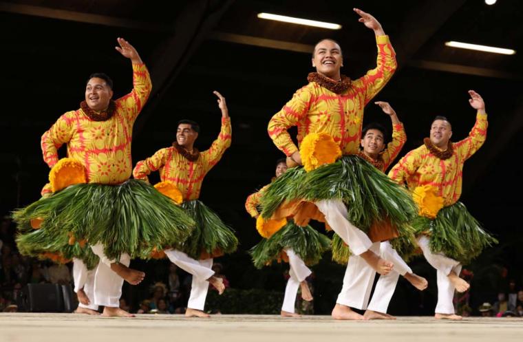 Group of hula performers dance in yellow patterned shirts and grass skirts holding gourd rattles