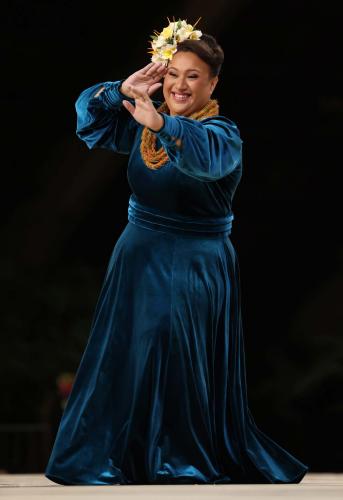 Woman performs hula on stage wearing a blue velvet dress and yellow floral headband
