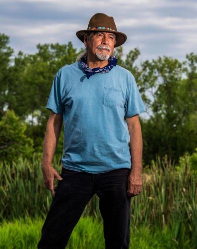 Daniel Wildcat stands in front of wetland wearing a wide brim hat and blue shirt