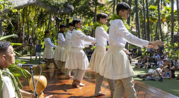 Group of boys in white shirts and white skirts perform hula to audience