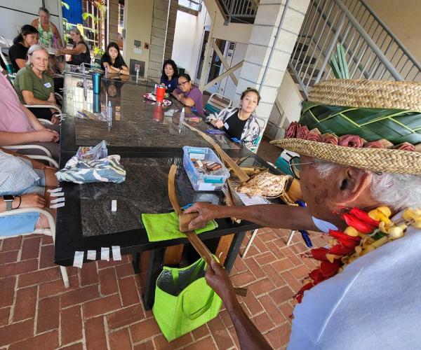 Man demonstrates weaving with hala leaves as workshop attendees watch around table