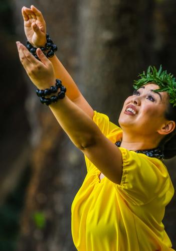 Person in yellow dress and green leaf crown reaches upward with both hands