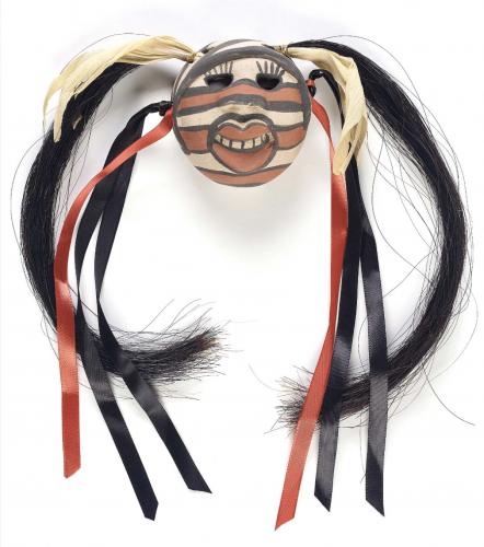 A smiling clay face decorated with white, black, and red stripes and two long tufts of horsehair