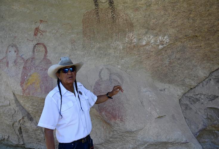 Person in uniform and white western hat points at paintings of people on a rock wall.