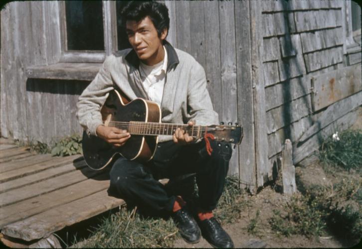 Man with pompadour and grey jacket sitting on porch playing an acoustic guitar