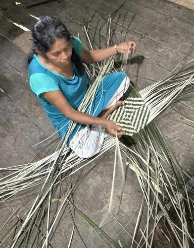 Weaver in blue dress sits on floor while weaving palms into a geometric pattern.