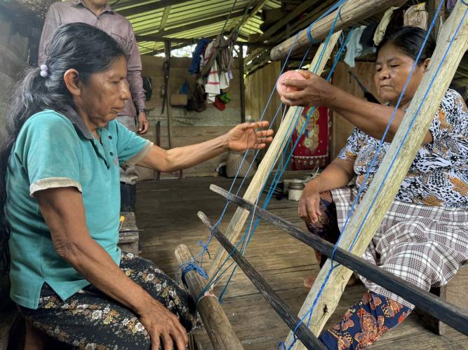 Two weavers pass yarn through a standing loom