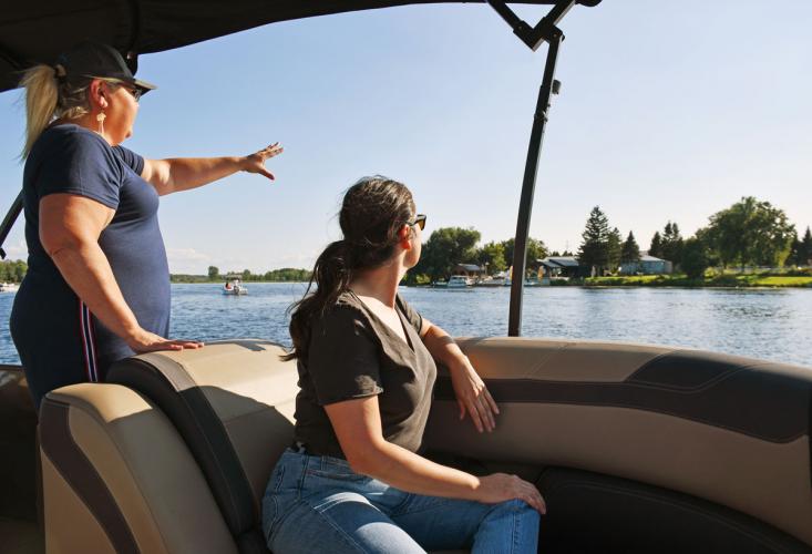 People in boat point at buildings on a treelined riverbank.