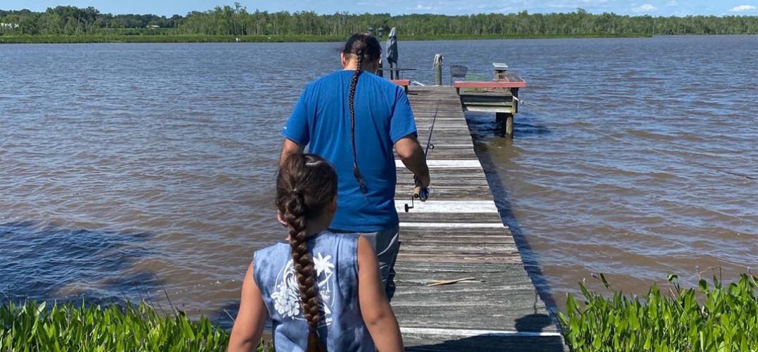 A father and son holding fishing poles walk down a dock toward a river