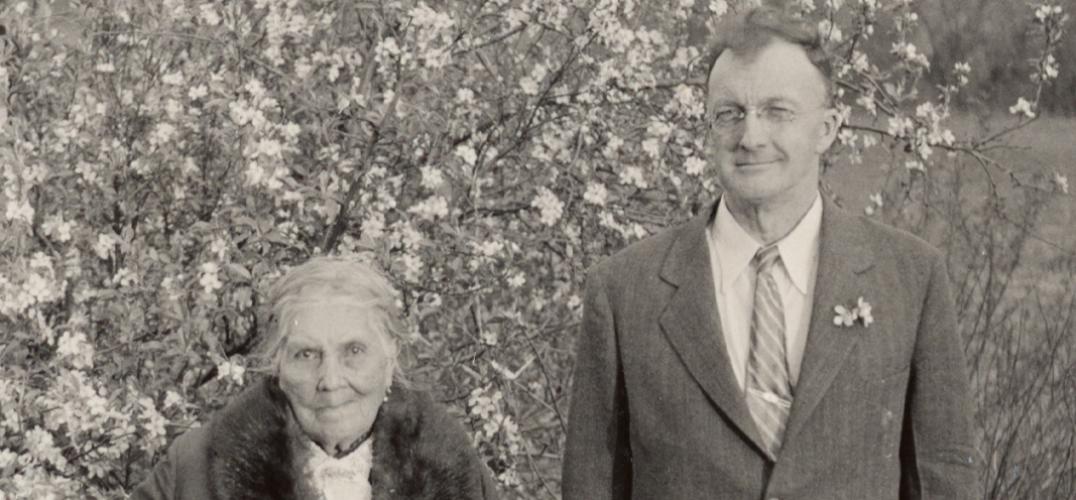 Isabel Meadows in a coat with a fur collar stands next to John Peabody Harrington wearing a suit in front of a tree