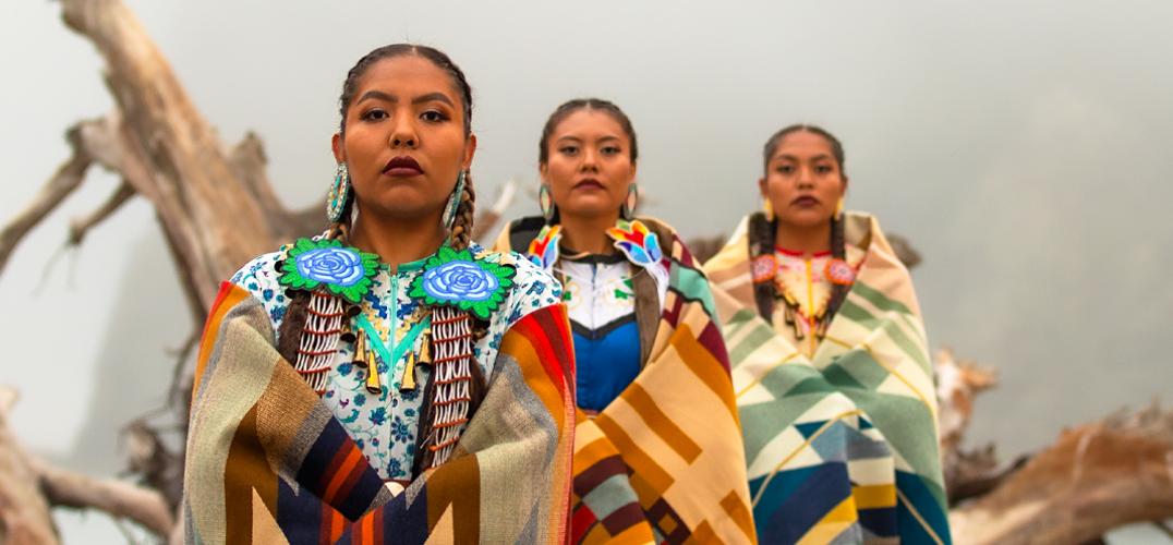 Three jingle dress dancers pose on a fallen tree log on the foggy shore.