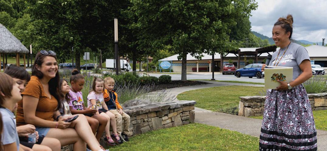A smiling woman holds a book, facing a semi-circle of children seated on a rock wall outdoors