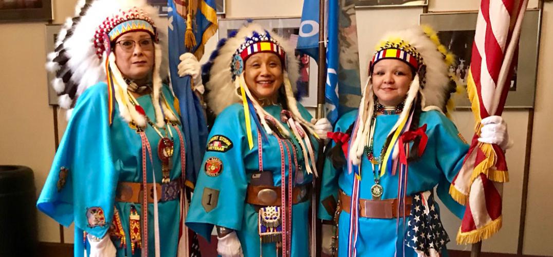 Three Native women veterans wearing blue traditional dresses with military decorations and and feather headdresses