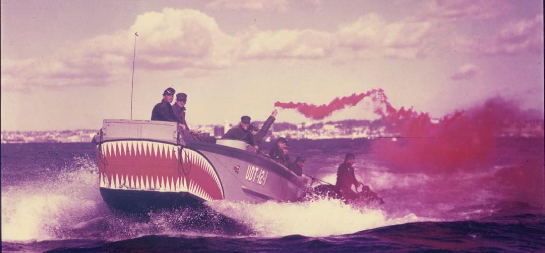 A 1950s photograph of a grey boat with teeth painted on one end carries 8 crew members on board. One crew member is holding a flare releasing red smoke.