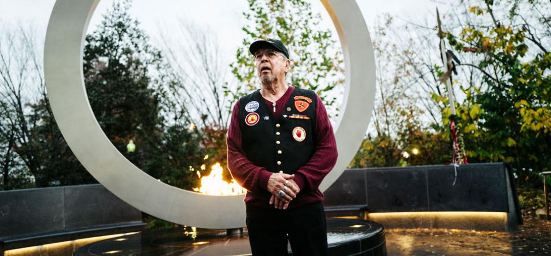A man in uniform stands in front of a tall metal circle with a lit flame at its base
