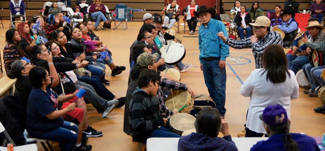 Crowd of people seated in a gymnasium observe participants playing a handgame. 