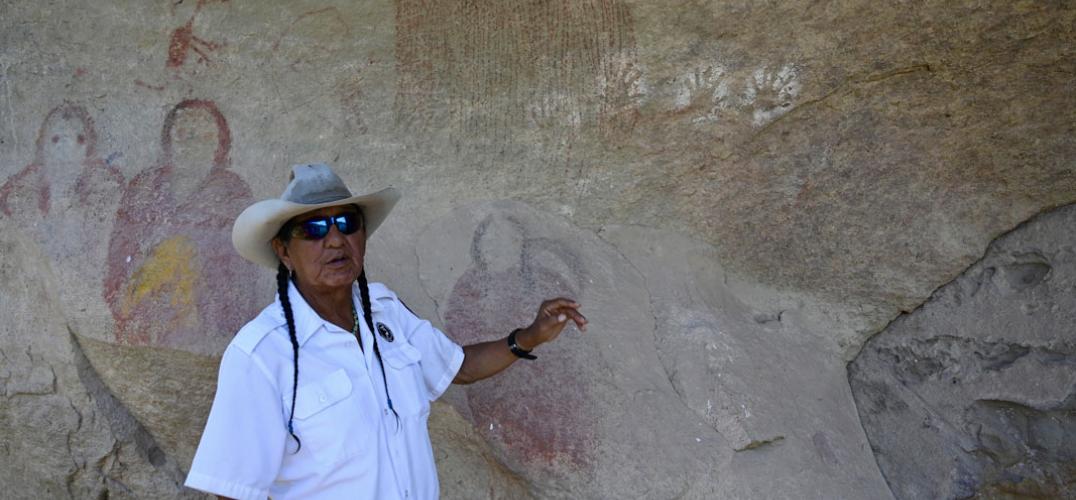 Person in uniform and white western hat points at paintings of people on a rock wall.