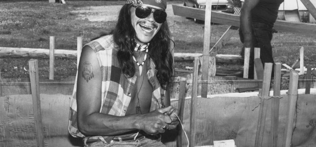 Person in aviator sunglasses, flannel vest, and bandana splits roots for a canoe-building workshop.