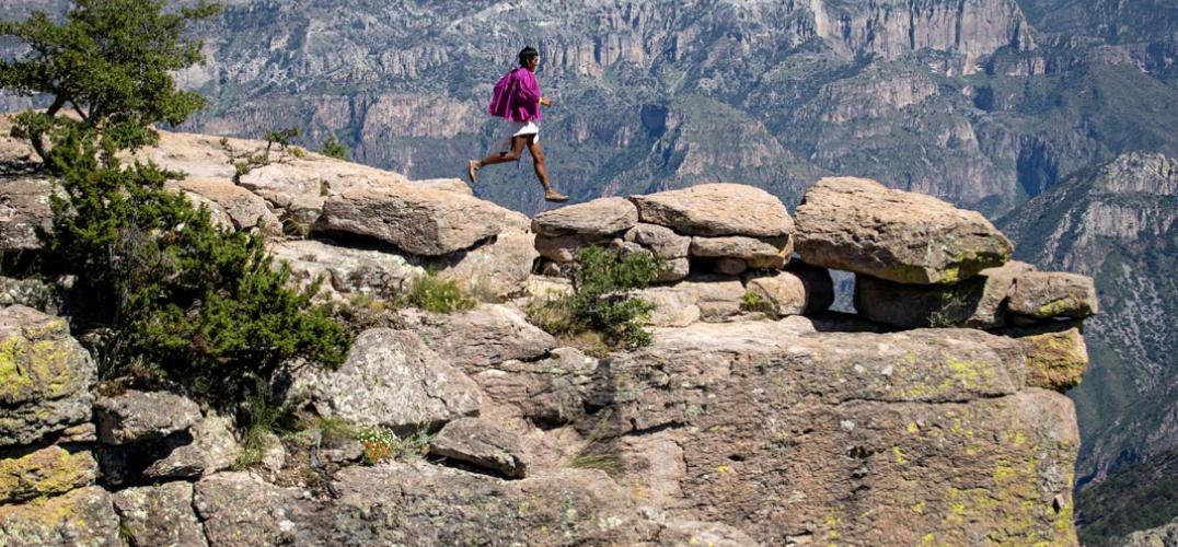 Person in traditional clothing and sandals running on top of rocky desert cliff.