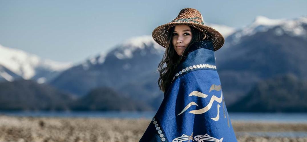 An Indigenous woman stands along a beach in a blue cloak depicting herrings.