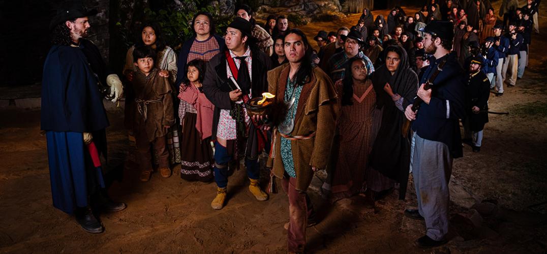 Actors on stage depict the Trail of Tears: a large group of Cherokee peoples are forcefully led by two soldiers.